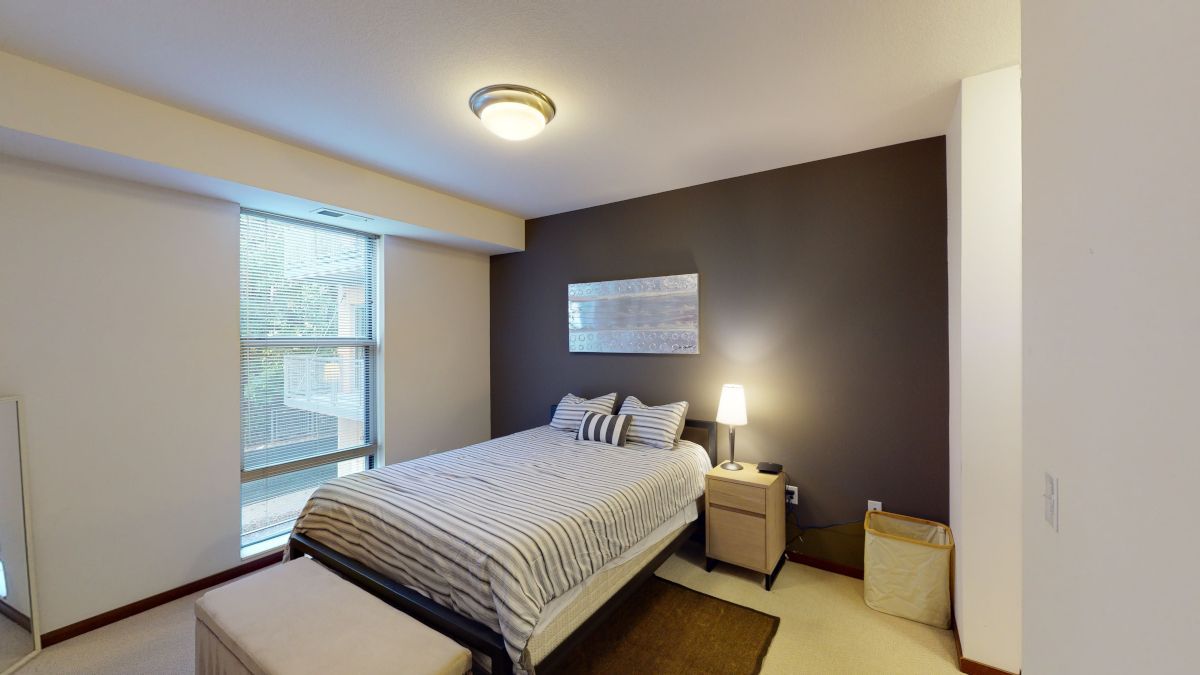 Small modern bedroom with a bed, nightstand lamp, window blinds, and dark accent wall.