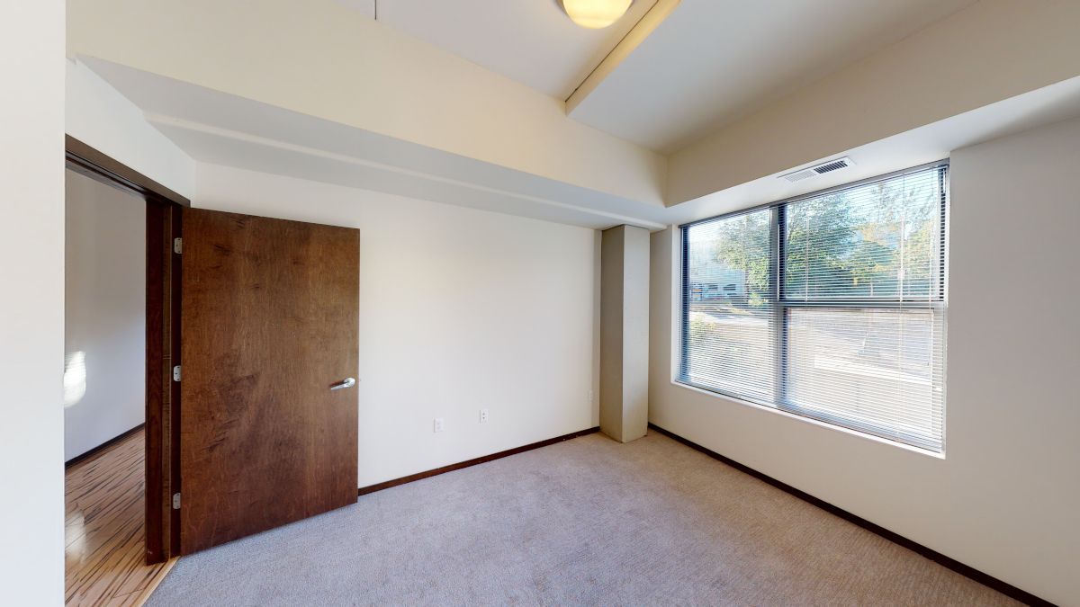 Empty bedroom with carpet, white walls, wooden closet doors, and a window with blinds