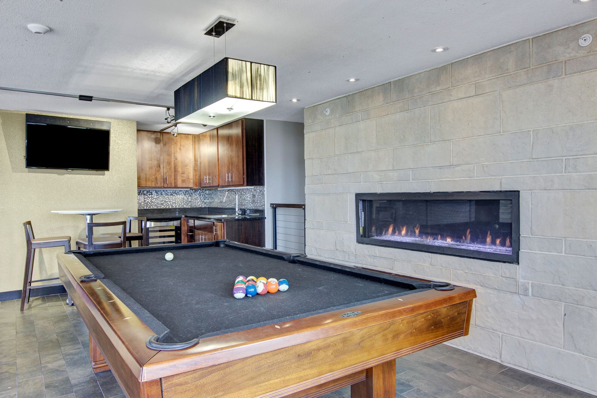 Modern game room with a pool table, fireplace, bar stools, and kitchenette.