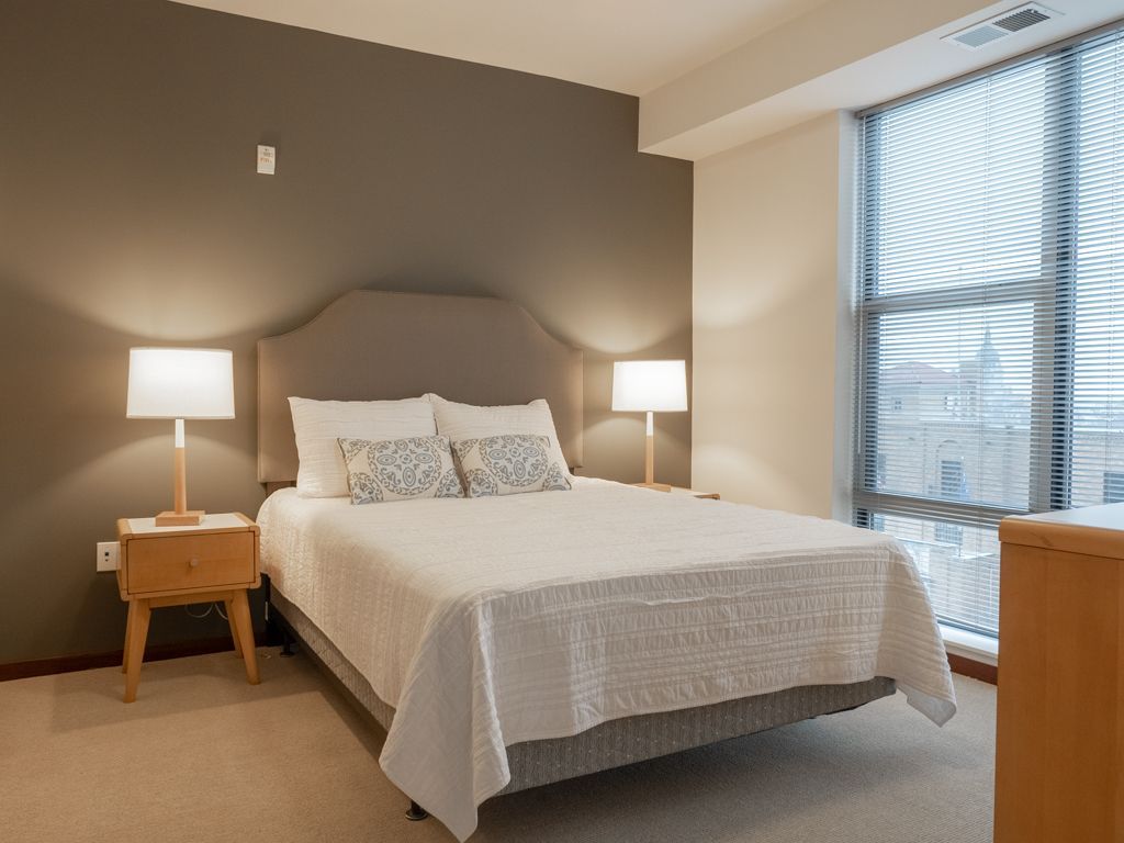 Modern bedroom with a made bed, bedside lamps, and a large window overlooking a city.