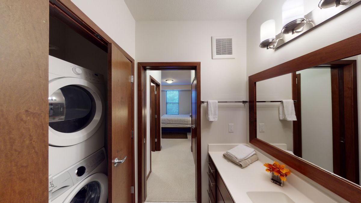 Narrow bathroom with stacked washer and dryer, mirror, sink, and doorway to bedroom