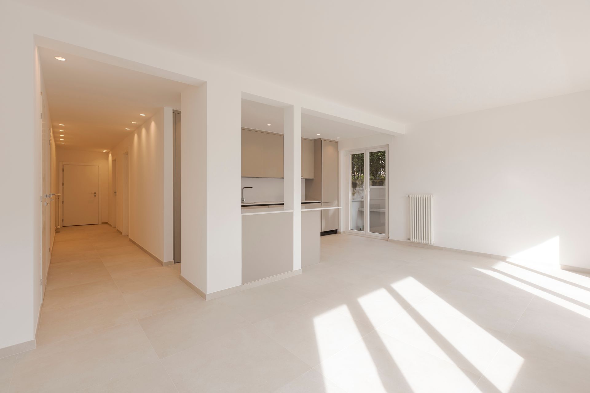 Empty, bright interior space with white walls, opening to a kitchen and hallway. Sunlight streams in from a doorway.