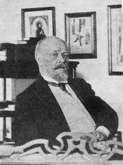 A formal, black-and-white portrait of a man with a beard and glasses sitting in a room with framed artwork on the wall.