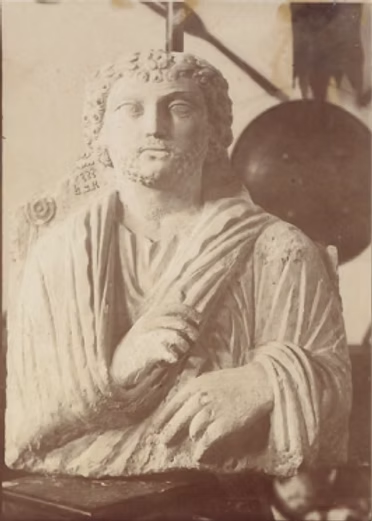 Sepia-toned historical photograph of a classical-style stone bust featuring a person draped in cloth, with a shield nearby.