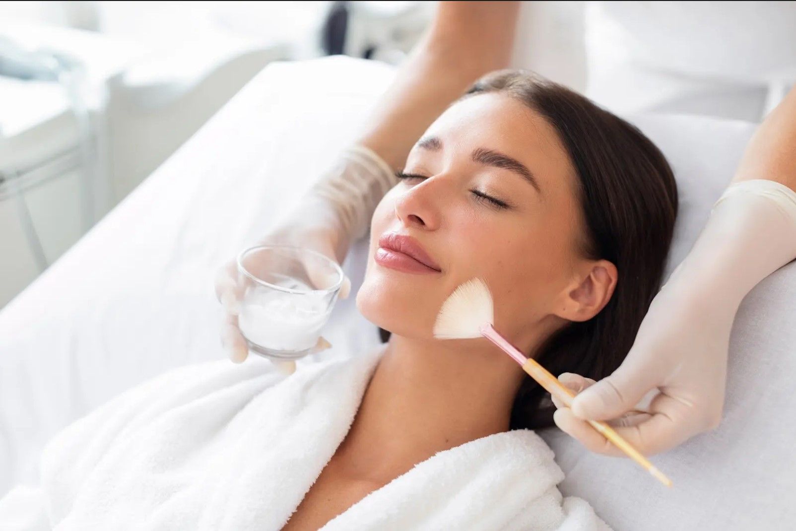 Woman receiving a facial treatment in a spa. A gloved esthetician applies a product with a brush.