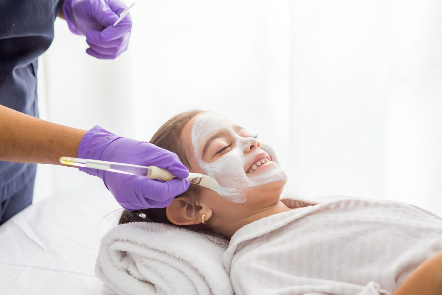 A child smiles while receiving a facial treatment in a spa setting; someone in purple gloves applies the mask.