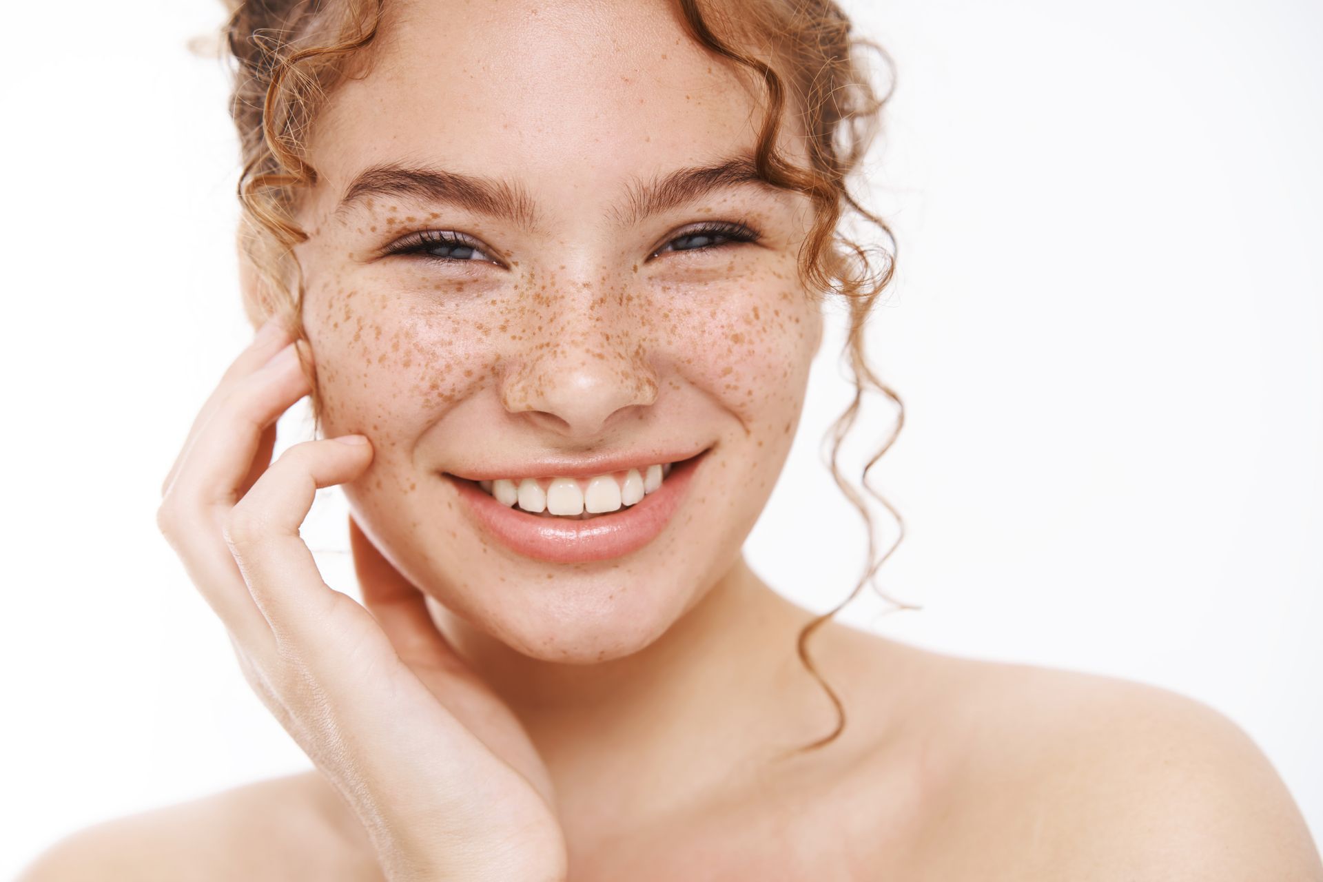 A woman with freckles on her face is smiling and touching her face.