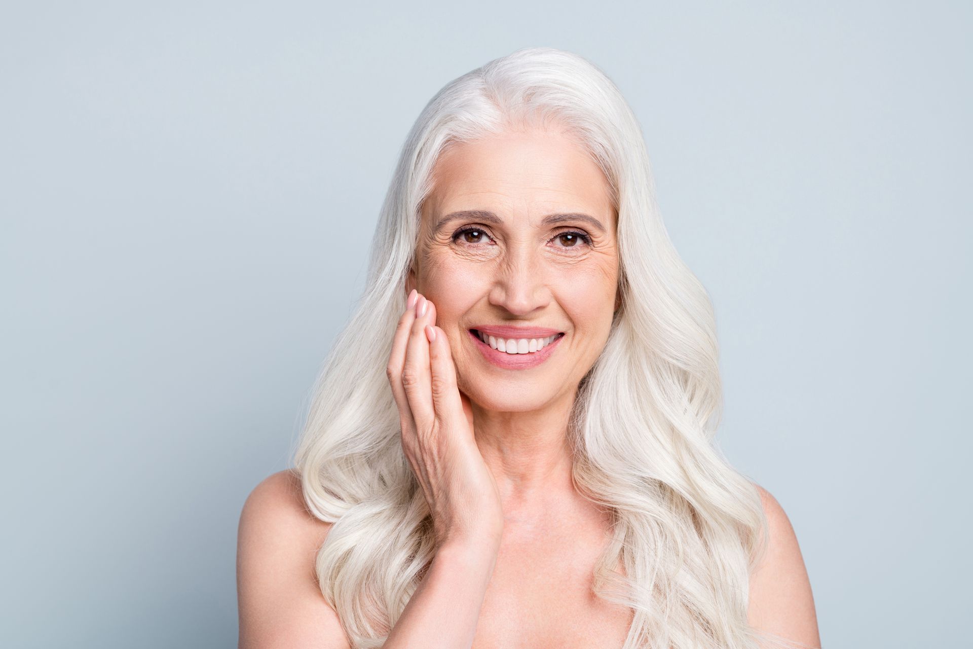 A woman with gray hair is smiling and touching her face.