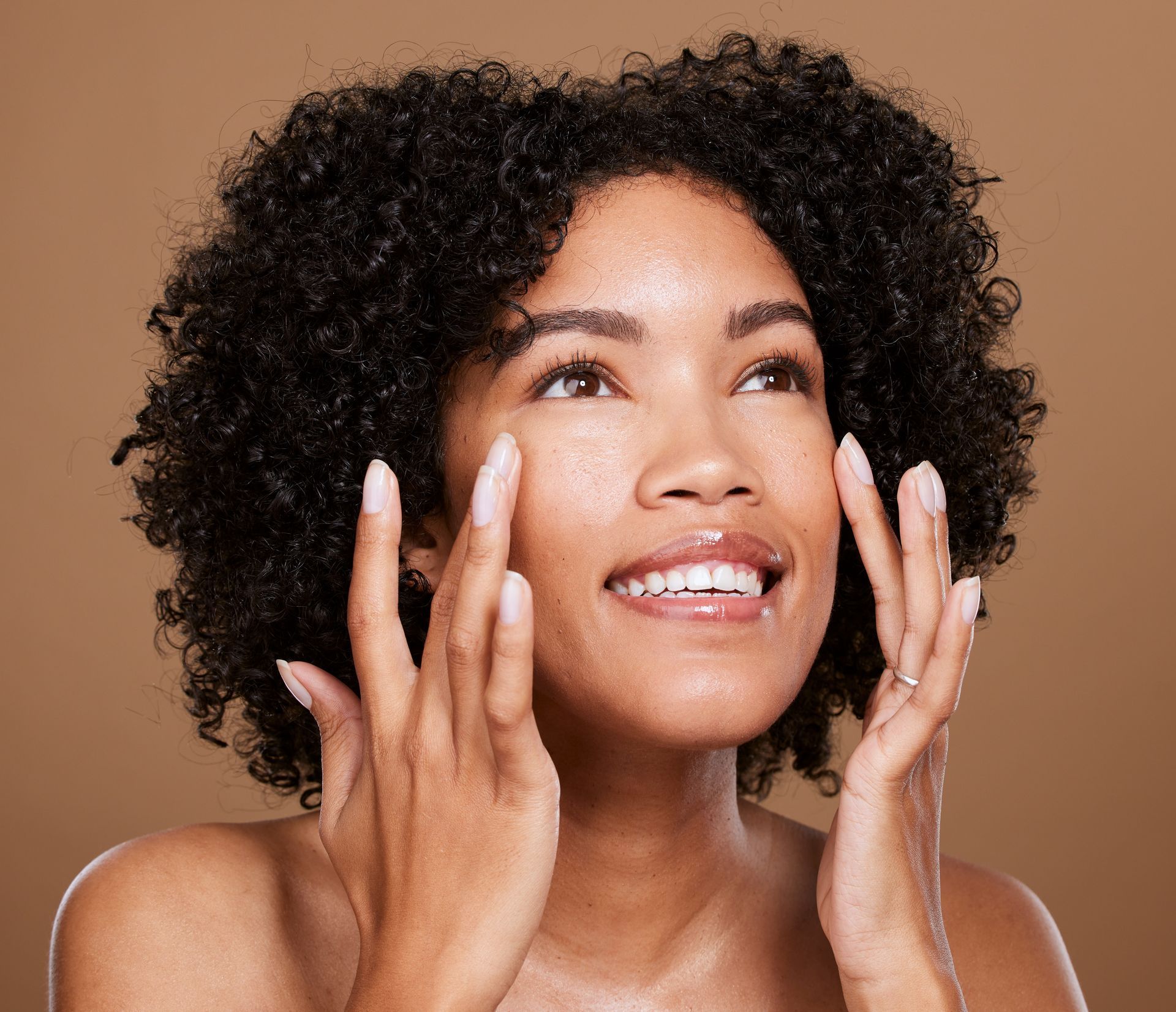 A woman with curly hair is smiling and touching her face.