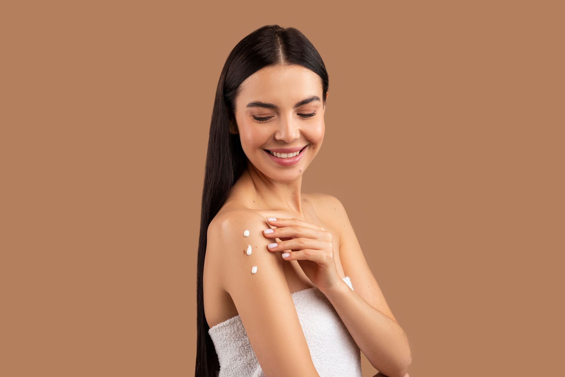 A woman wrapped in a towel is applying lotion to her shoulder.