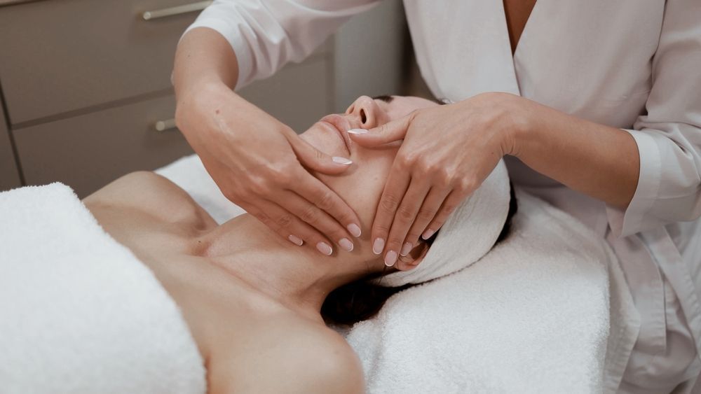Woman receiving facial massage at a spa, hands on face, draped in white, serene expression.