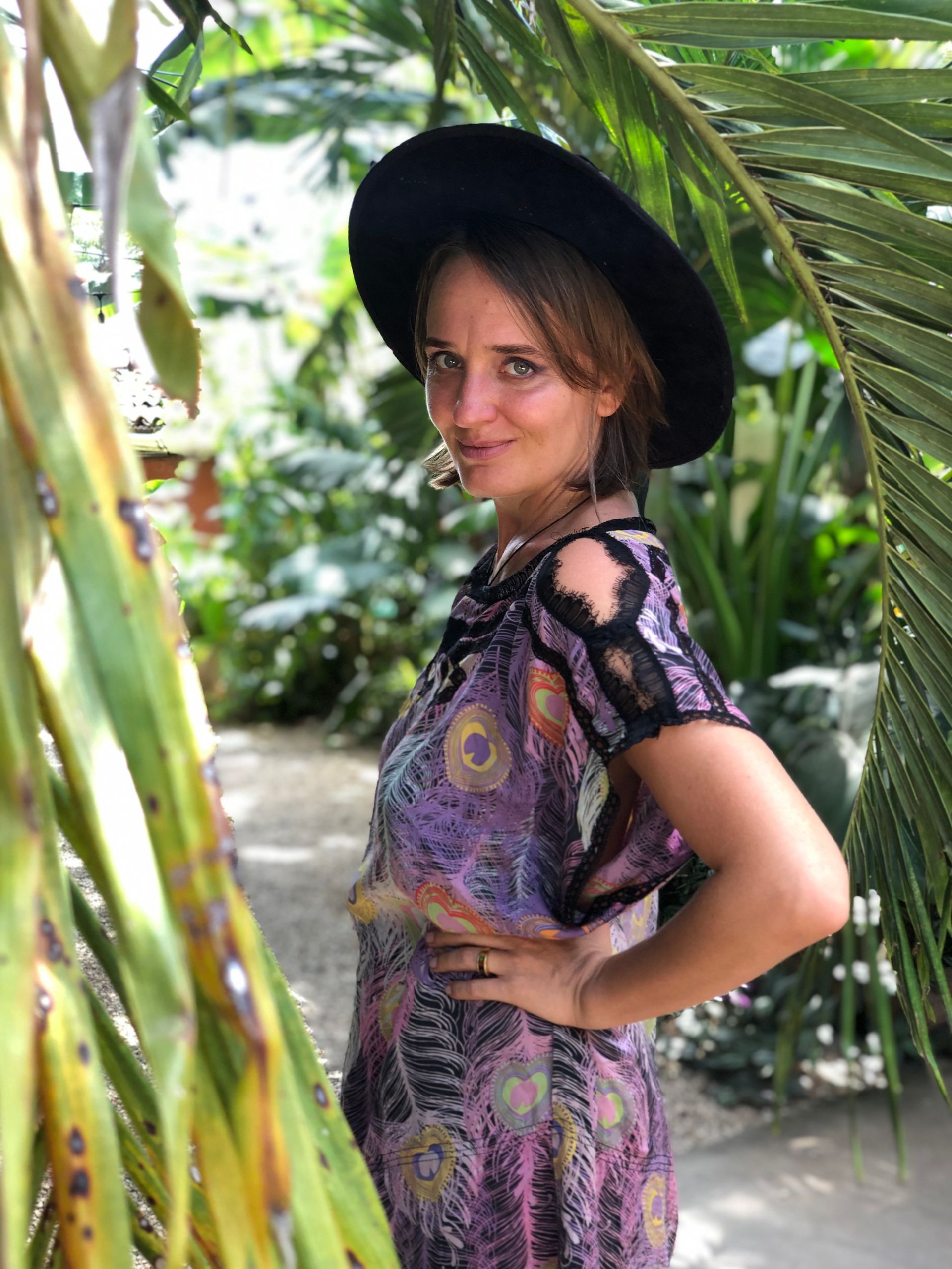 A woman wearing a black hat and a purple dress is standing in front of a palm tree. Jane Stark