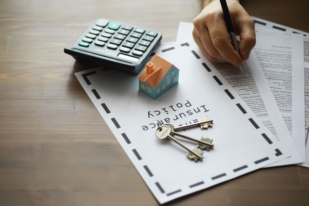 Person writing on insurance policy with house model, keys, and calculator on a wooden table.