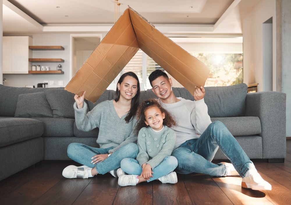 Family of three under cardboard roof, smiling in living room.