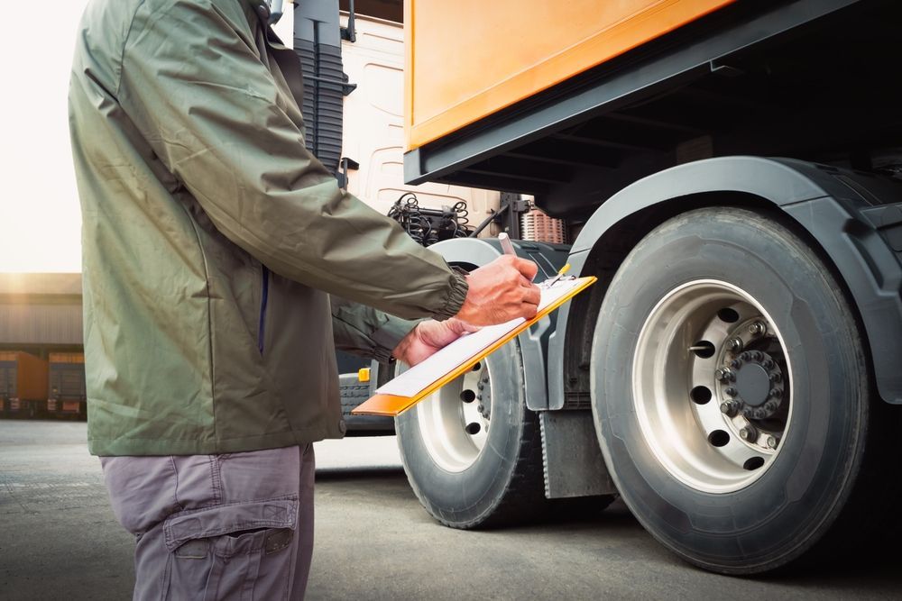 Person in green jacket inspecting semi-truck tires, holding clipboard.