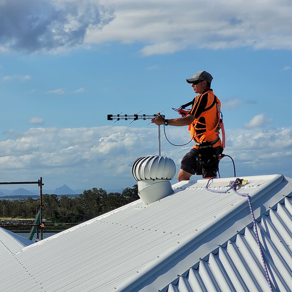 A Man Is Standing On Top Of A Roof Holding An Antenna — Brisbane Antenna Specialists in Burpengary East, QLD