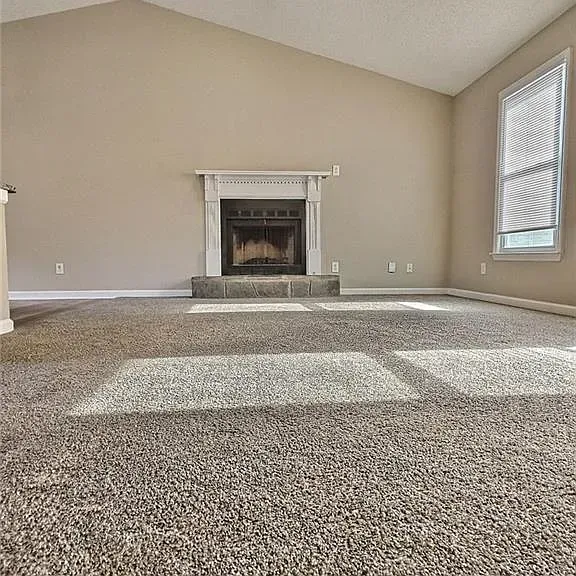 Empty living room with a fireplace and window; neutral-toned walls and carpet.