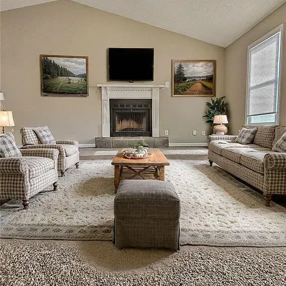 Cozy living room with fireplace, TV, armchairs, sofa, rug, art, and ottoman. Neutral colors, natural light.