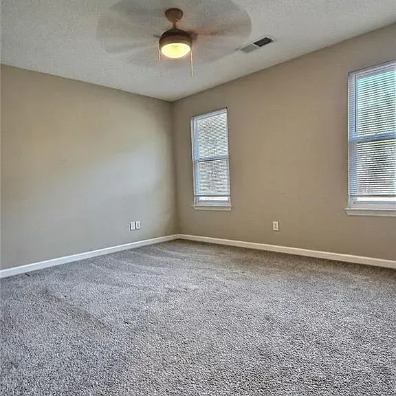 Empty room with gray carpet, beige walls, two windows, and a ceiling fan.