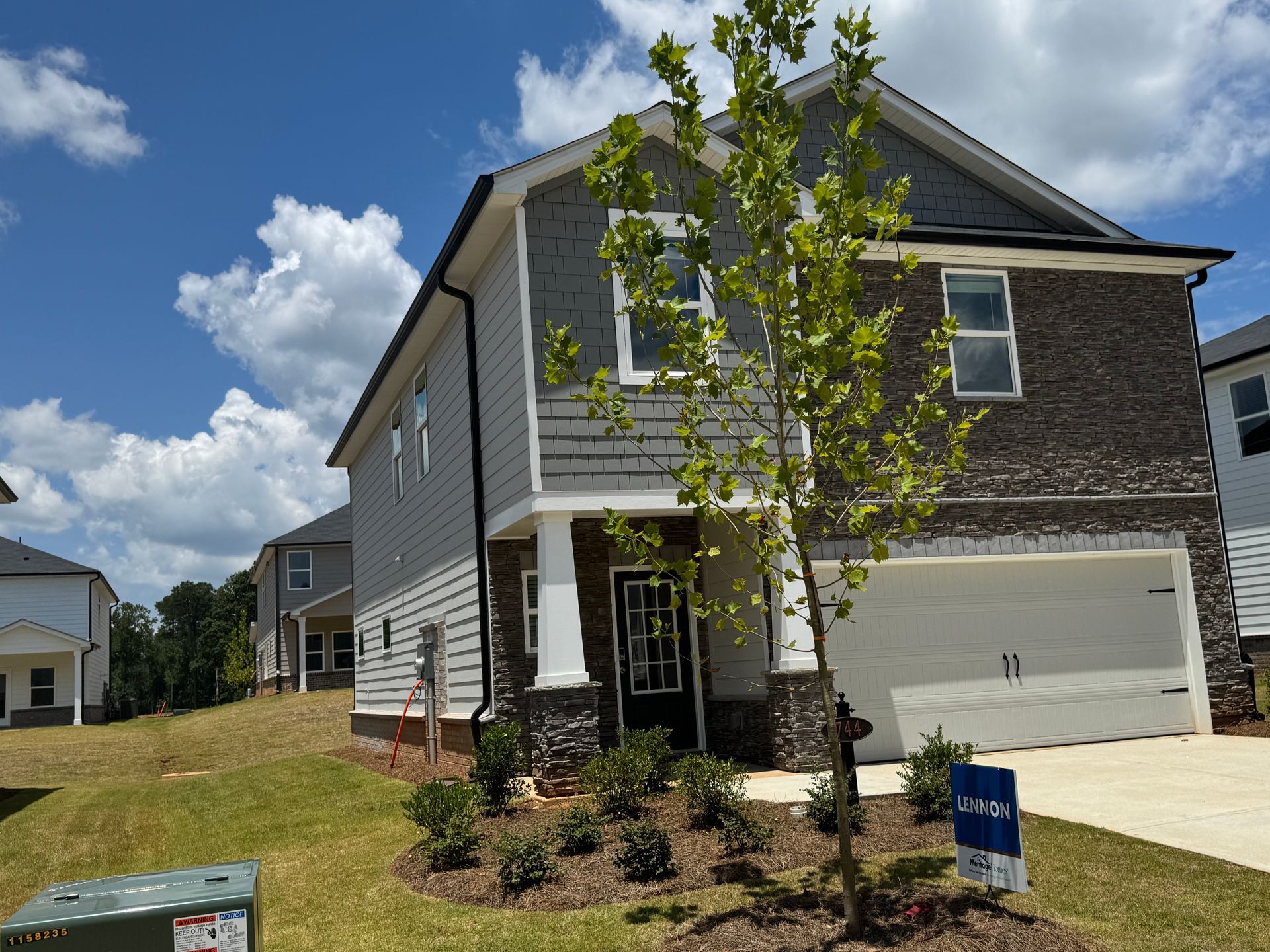 Two-story house with gray siding and brick facade; front yard with newly planted shrubs and a small tree.