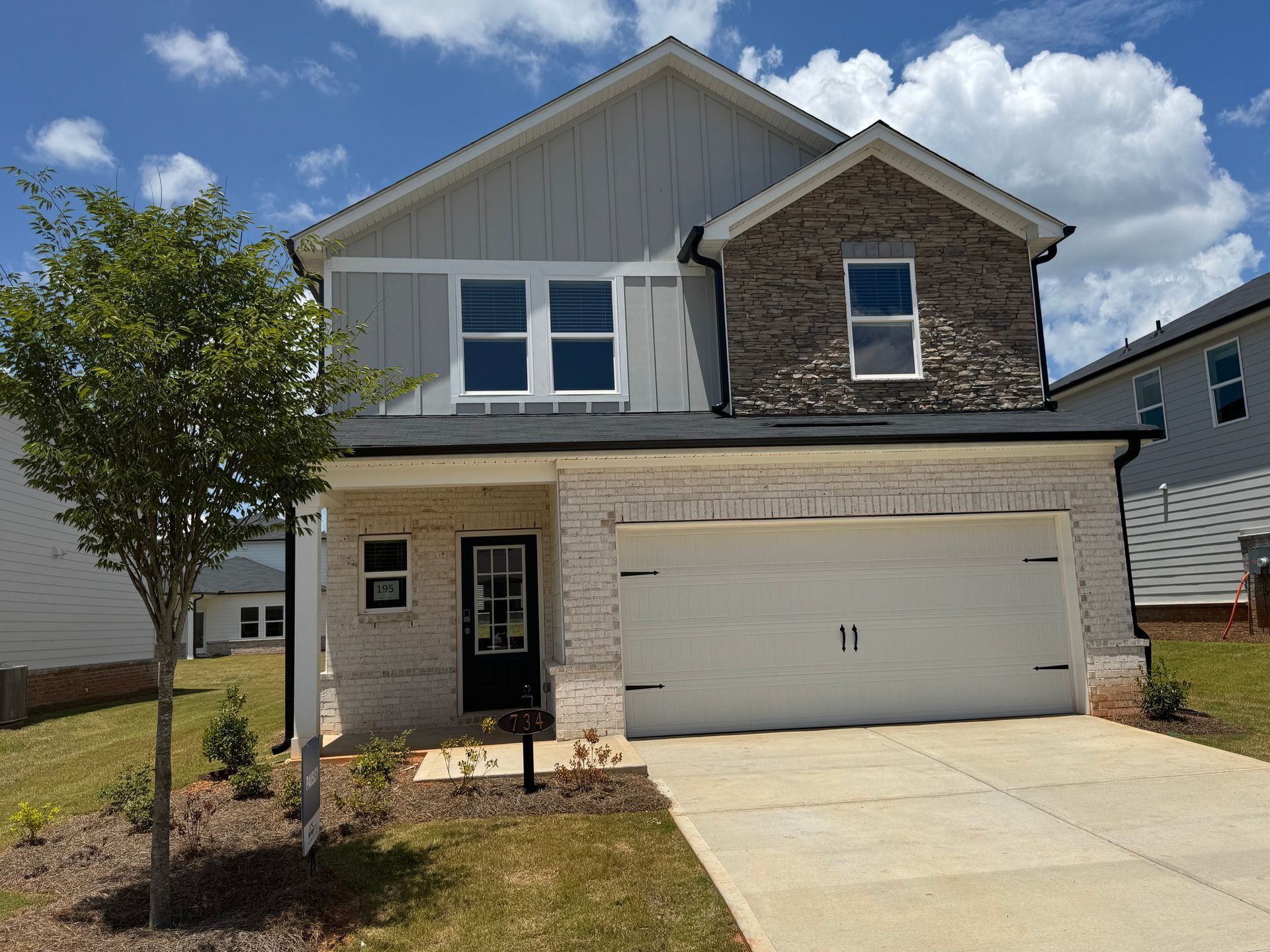 Two-story house with gray siding, brick facade, and a white garage door, set on a grassy lot under a blue sky.