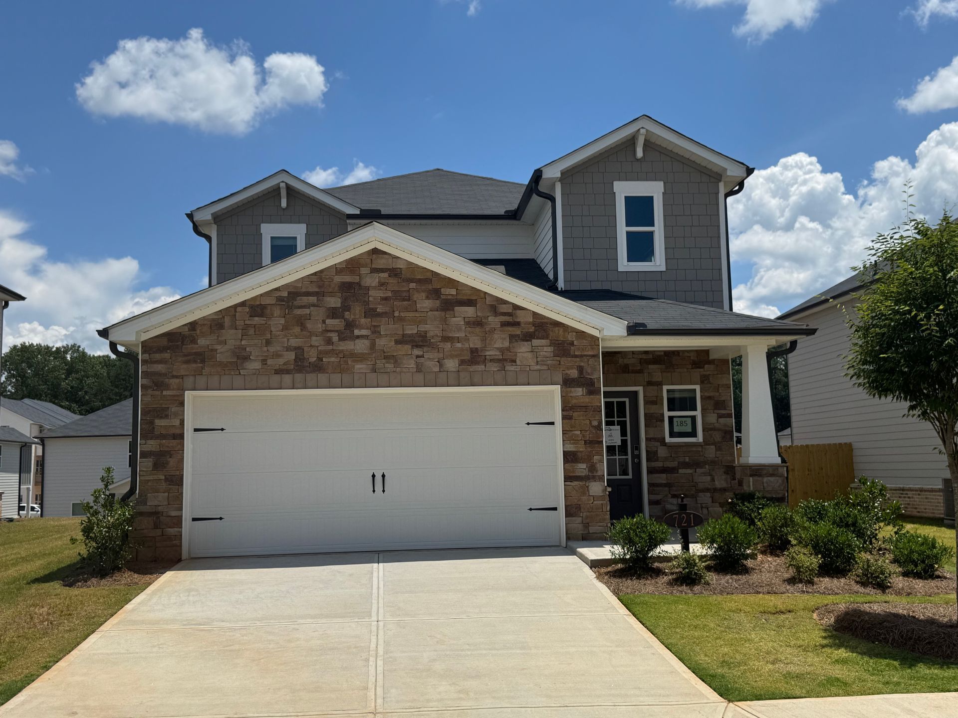 Two-story house with gray siding, brick facade, white garage door, and concrete driveway under a blue sky.