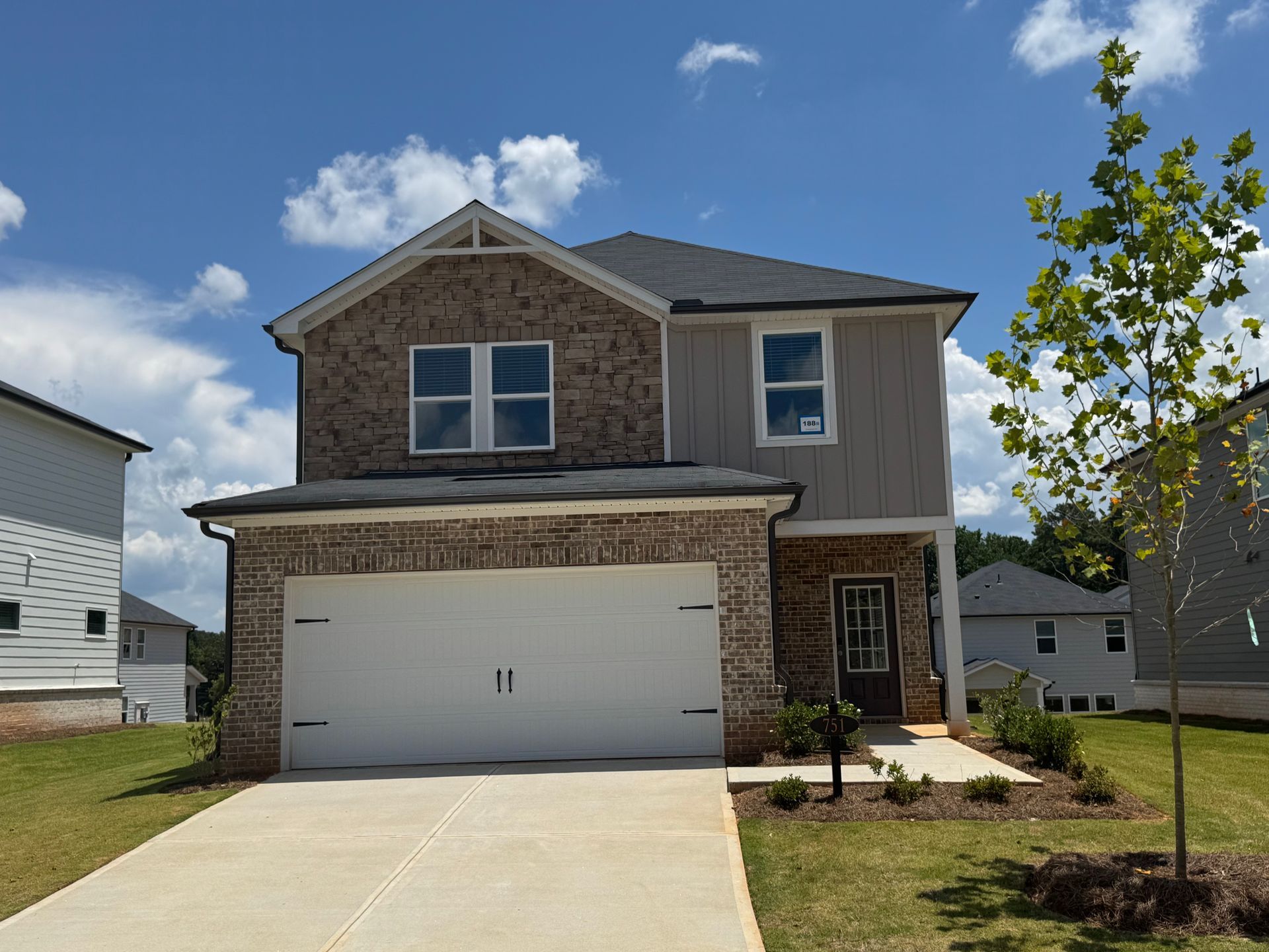 Two-story house with brick and gray siding, white garage door, and a small tree in the yard.