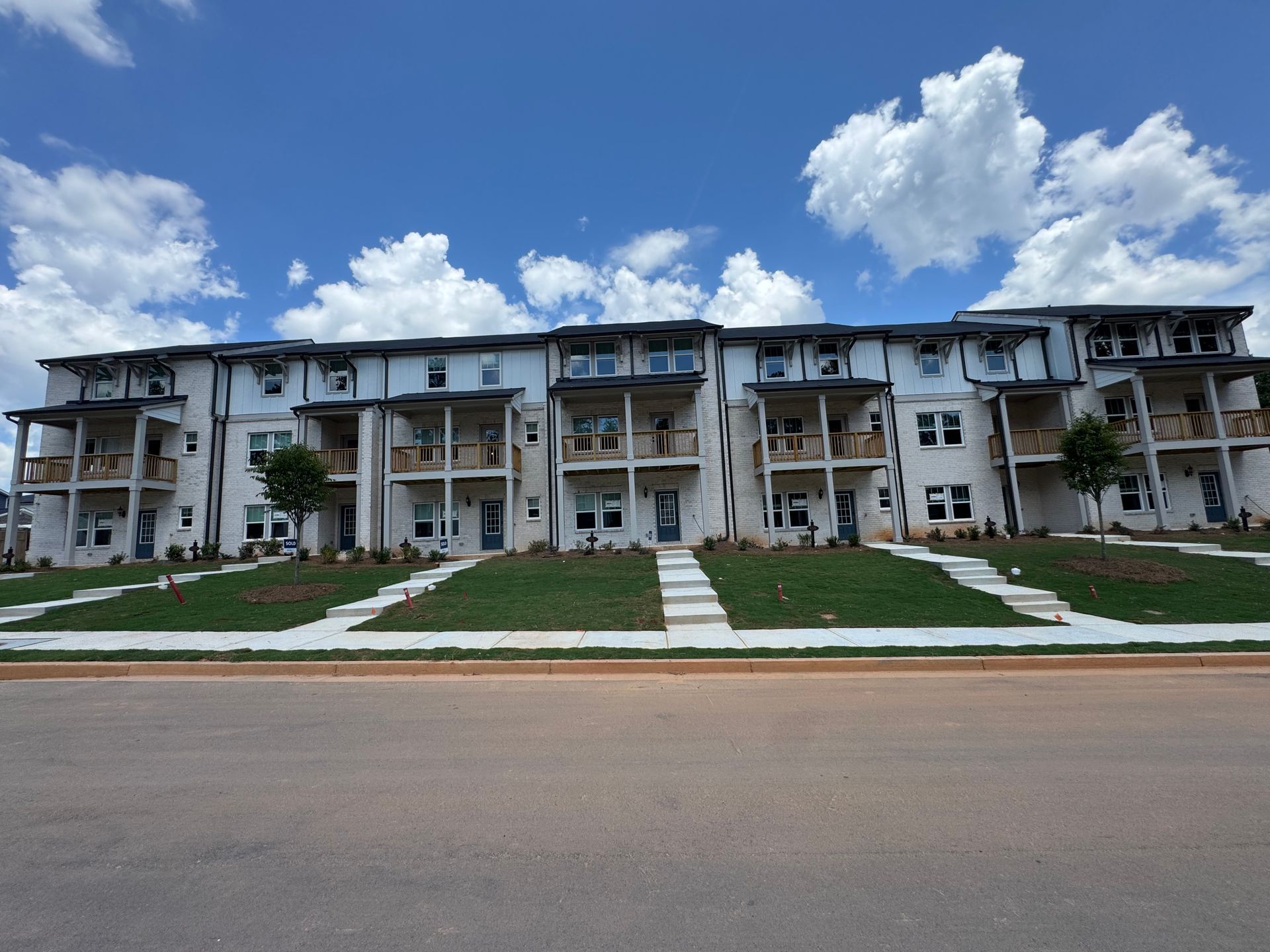 Row of white townhomes with balconies, set on a grassy hill with a blue sky and clouds overhead.