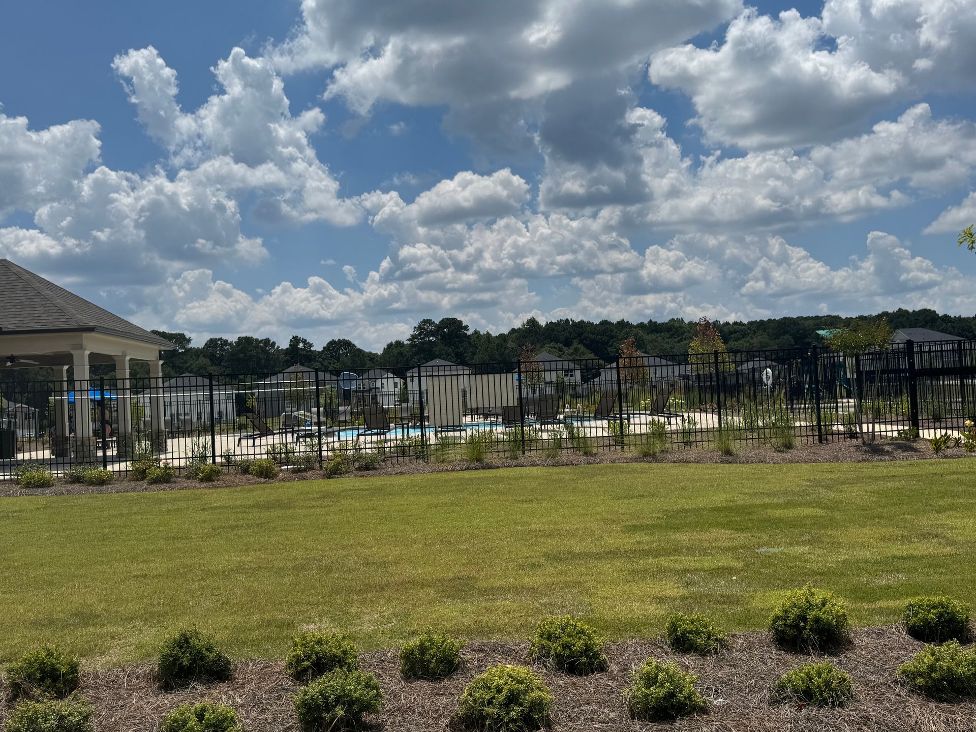 Green lawn leads to a fenced pool area with houses and a gazebo under a cloudy sky.