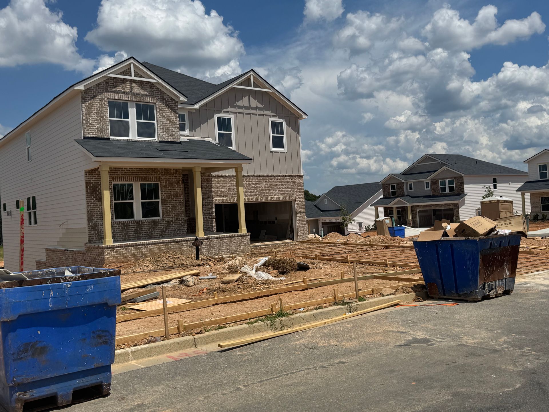 New houses under construction; one with tan siding, brick, and a porch, two blue dumpsters in foreground.