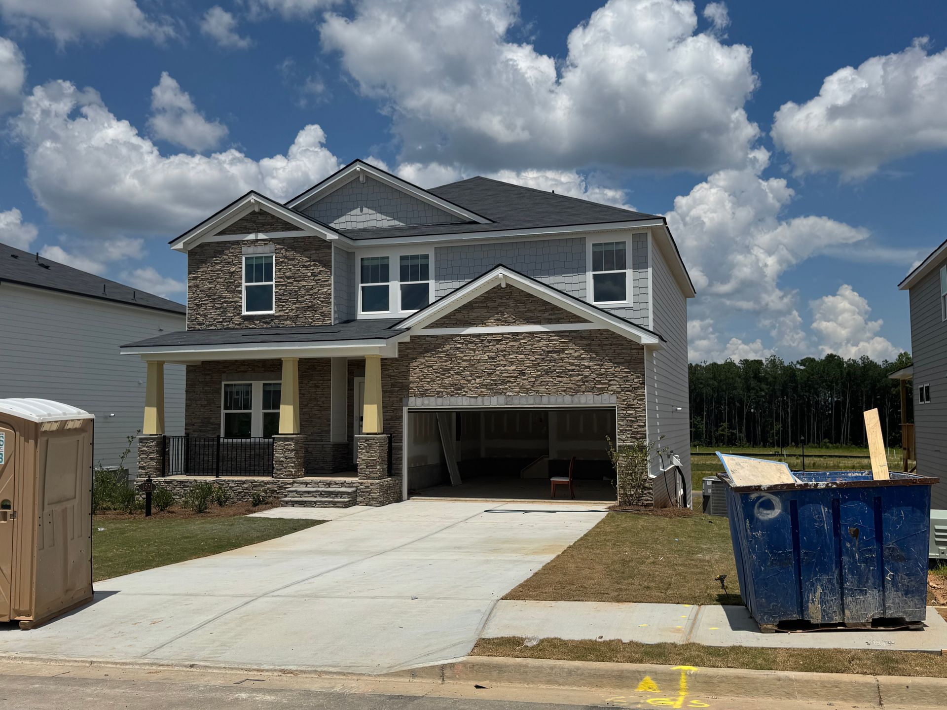 Two-story house under construction, with stone facade accents, blue sky, and a porta-potty in front.