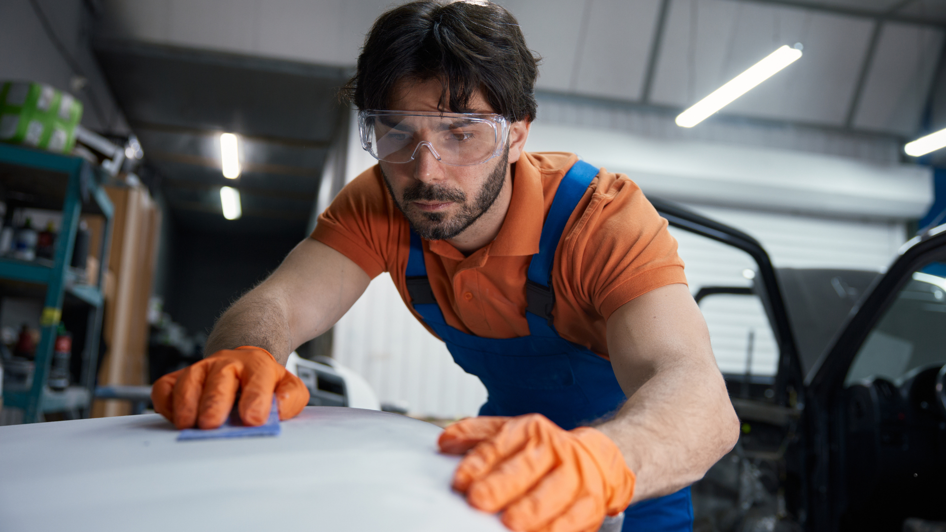 A technician in orange gloves and protective eyewear meticulously sanding the surface of a car as part of the auto reconditioning process.