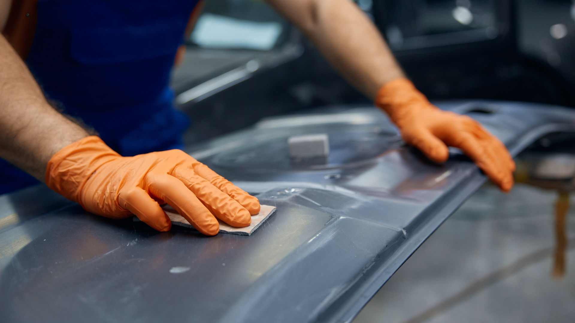 A technician in orange gloves using a sanding block on a vehicle's surface, performing a detail-orie
