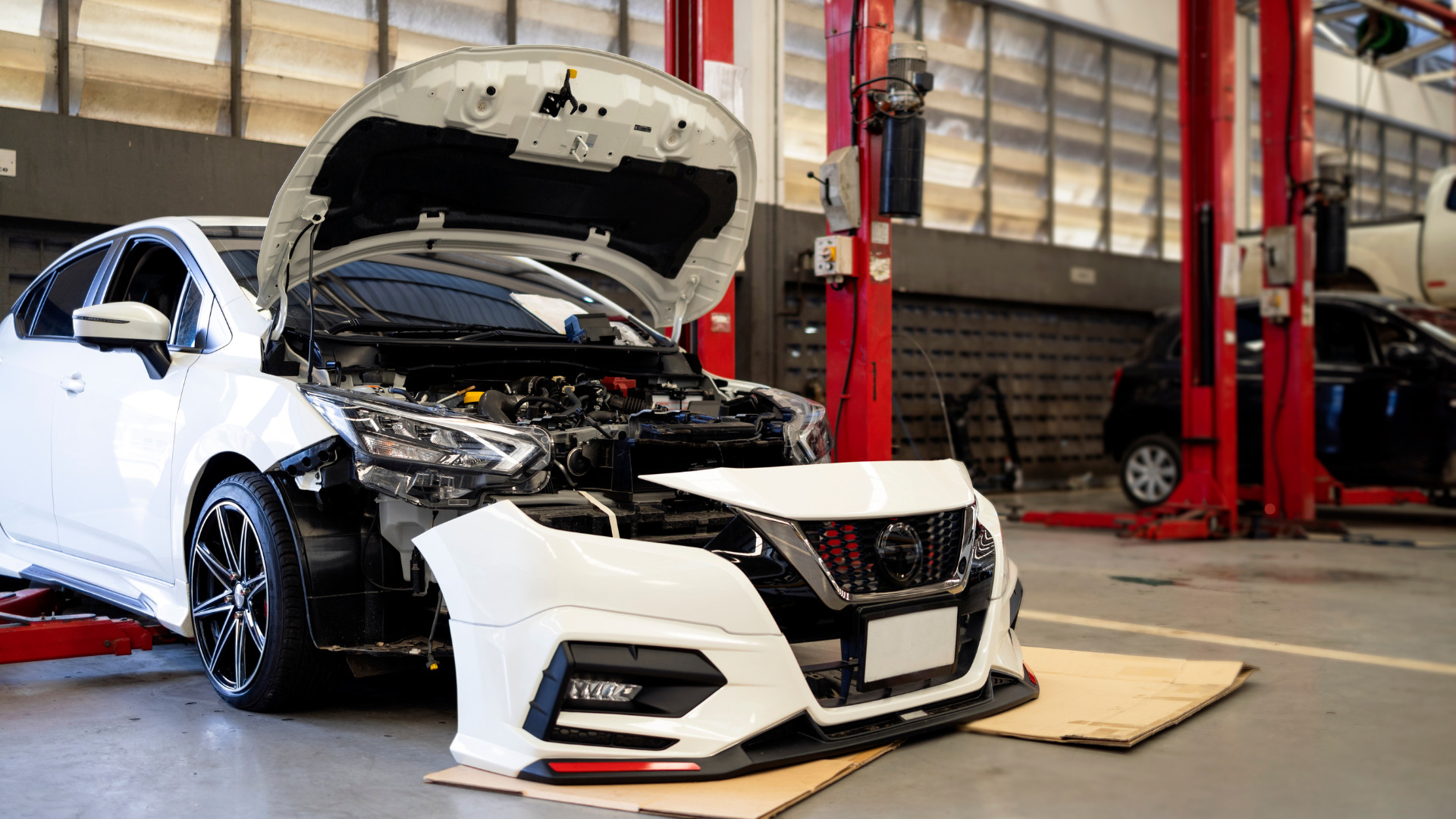 A white Nissan car with the front bumper removed, lifted on a repair shop lift with its hood open, s