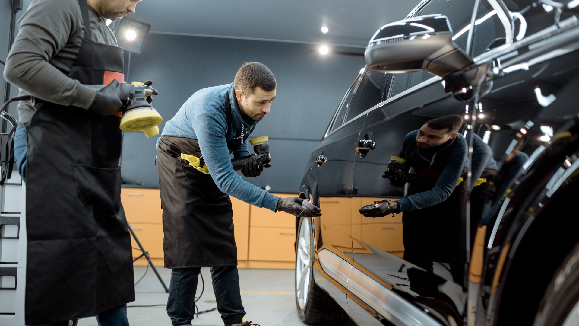 Two technicians in aprons detail the glossy black exterior of a car in a professional, well-lit studio.