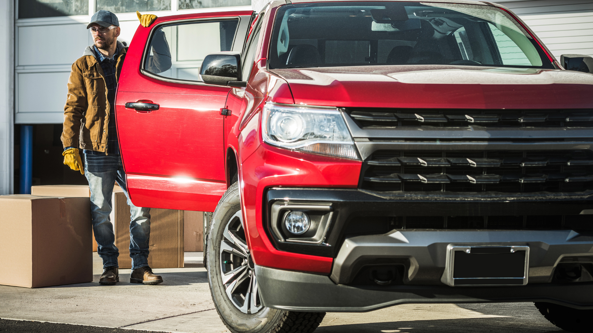 A man wearing a brown jacket and yellow gloves opening the driver-side door of a red pickup truck ou