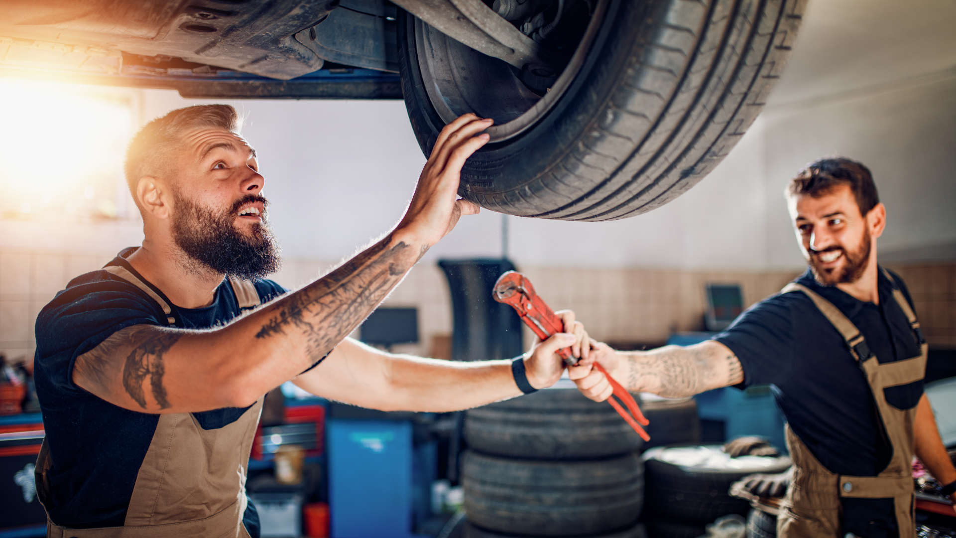 Two auto mechanics working together in a workshop, one holding a tire and the other using a wrench t