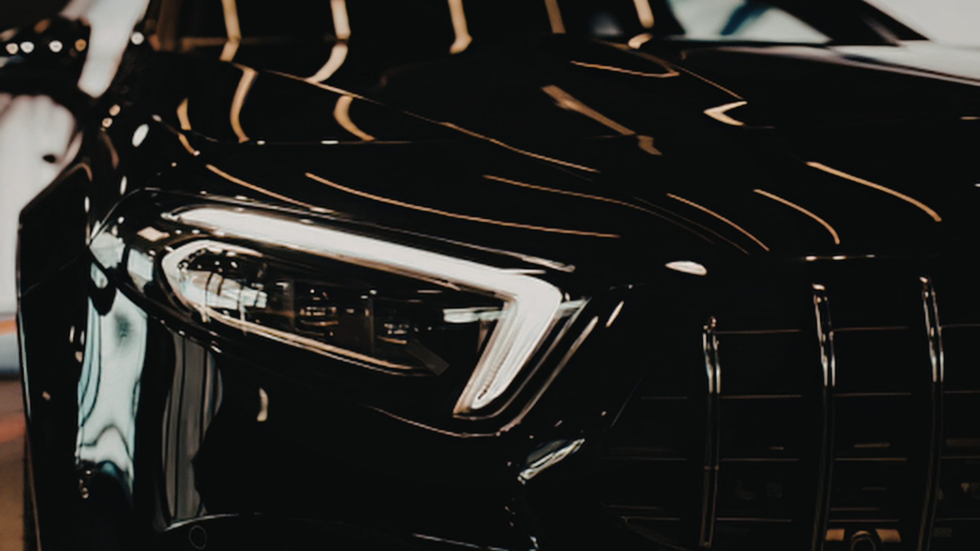 Close-up shot of the sleek front of a black luxury car with its headlight shining brightly under a well-lit environment.