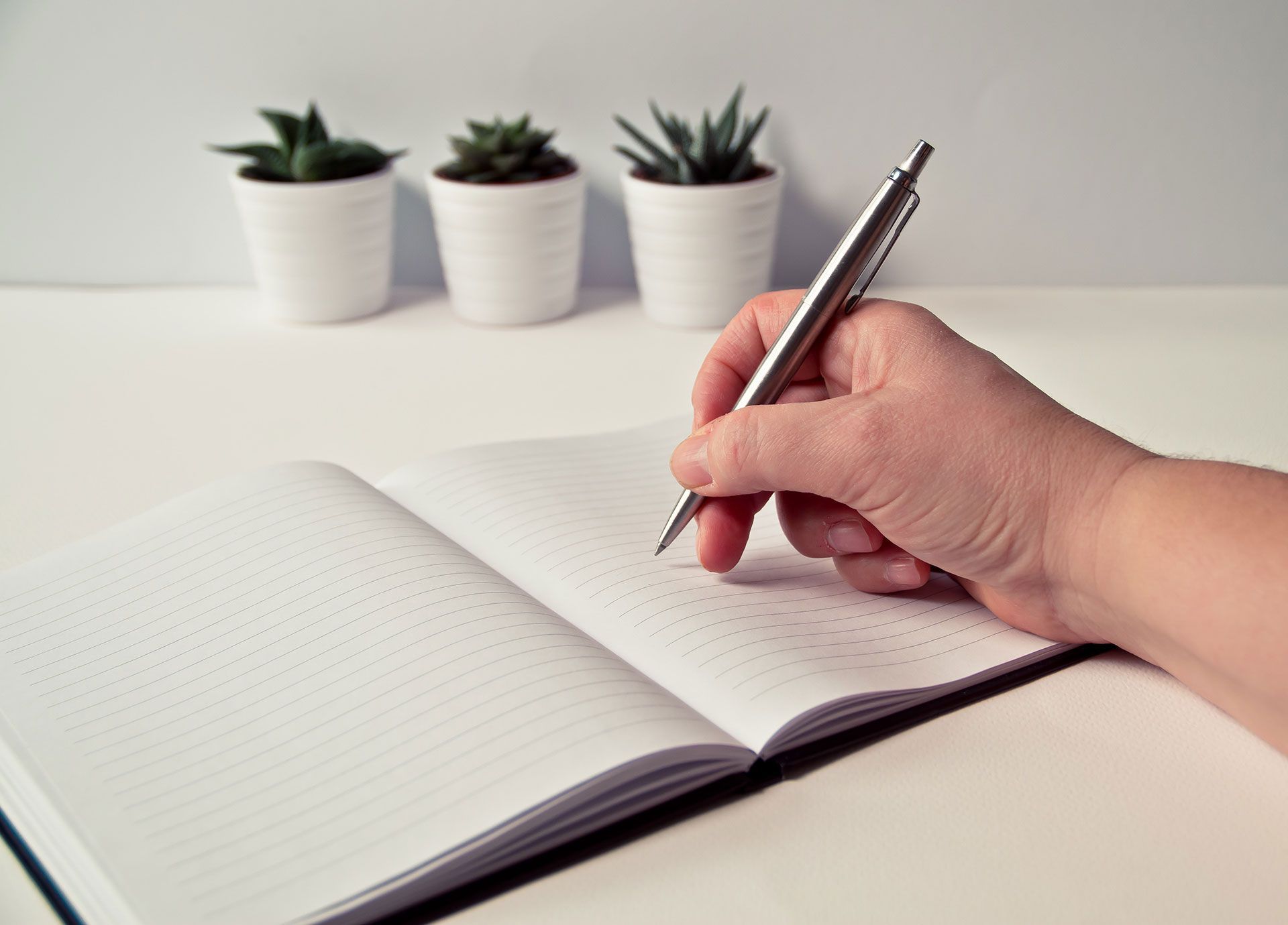 Hand writing in open notebook with pen on white desk; small potted plants in background.