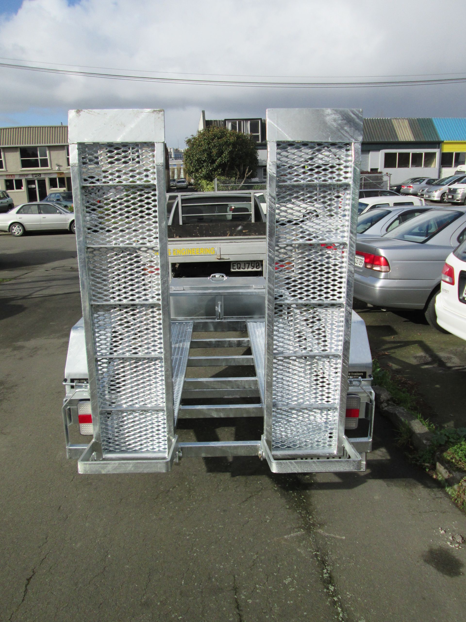 Trailer with two grated ramps, viewed from the rear, parked on pavement. Background includes cars and buildings.