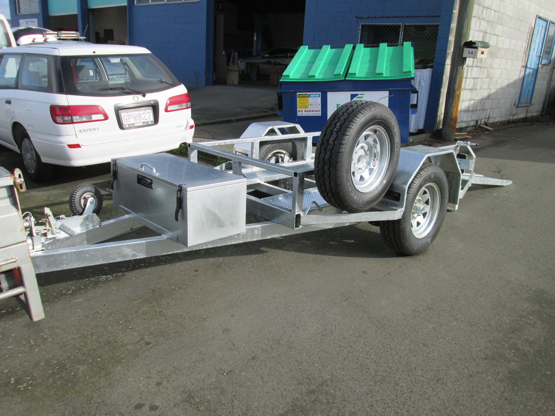 Silver utility trailer with a spare tire, parked on pavement next to a white car.