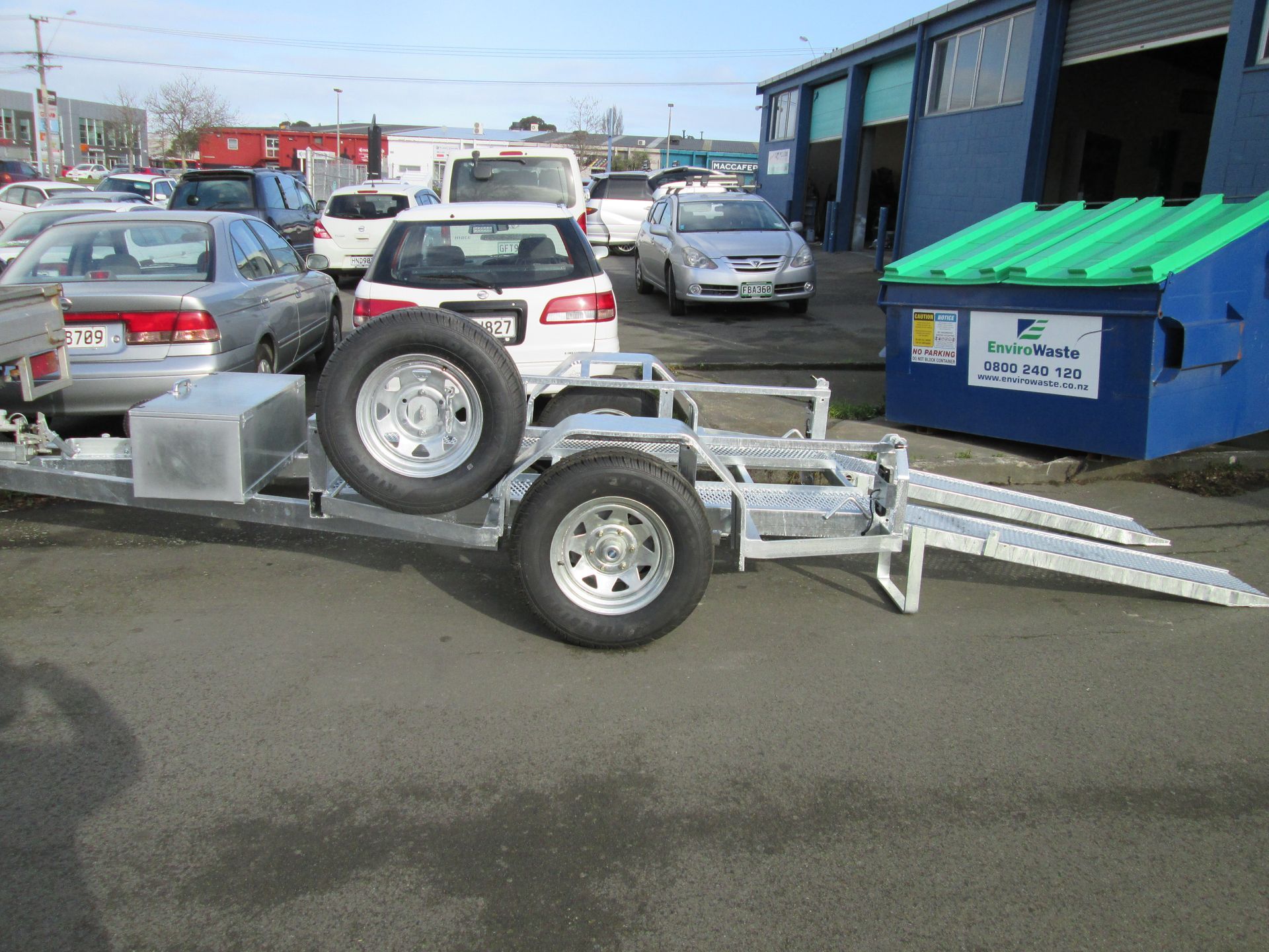 Trailer with two wheels, spare tire, and loading ramps parked outside a building.