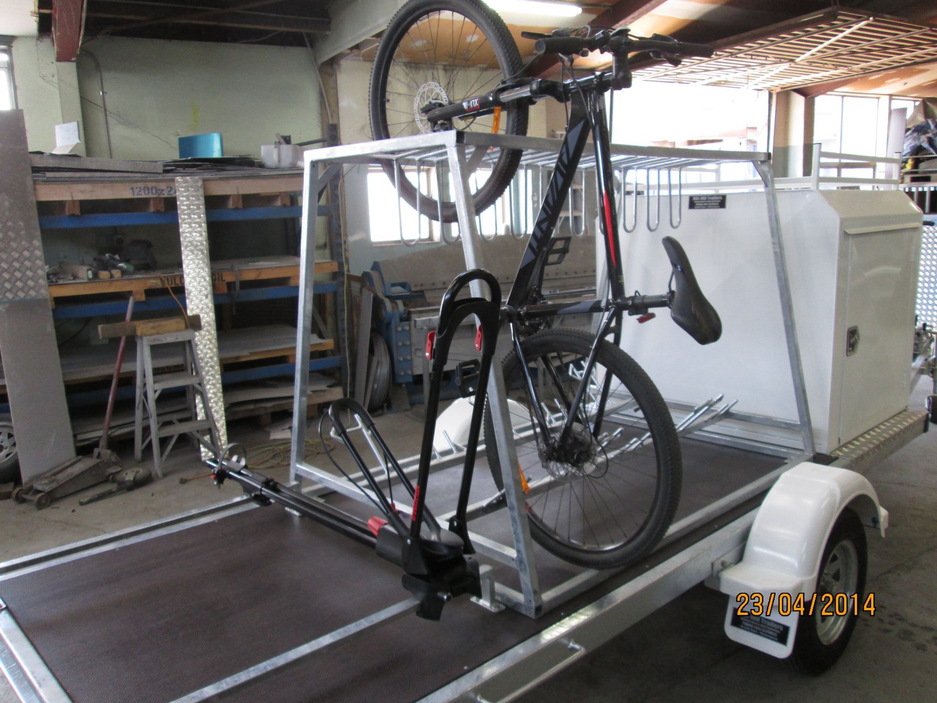 Bike secured on a trailer with metal frame, ready for transport. White trailer, black bike. Workshop background.