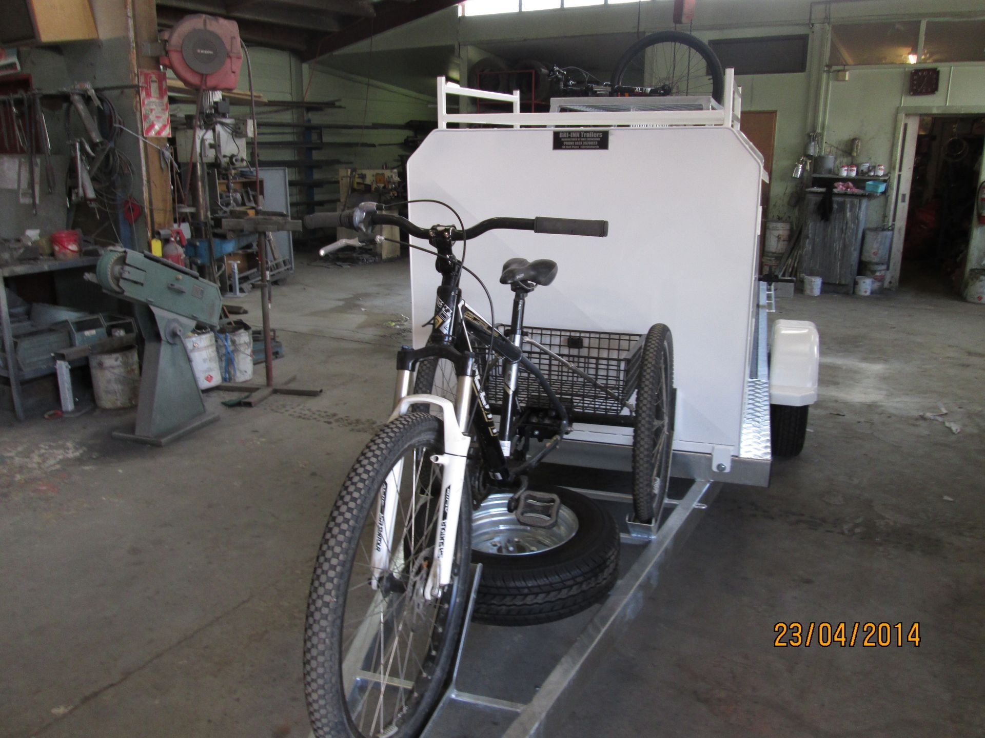 Tricycle and bicycle on a small trailer inside a workshop. Gray metal, white trailer, sunny.