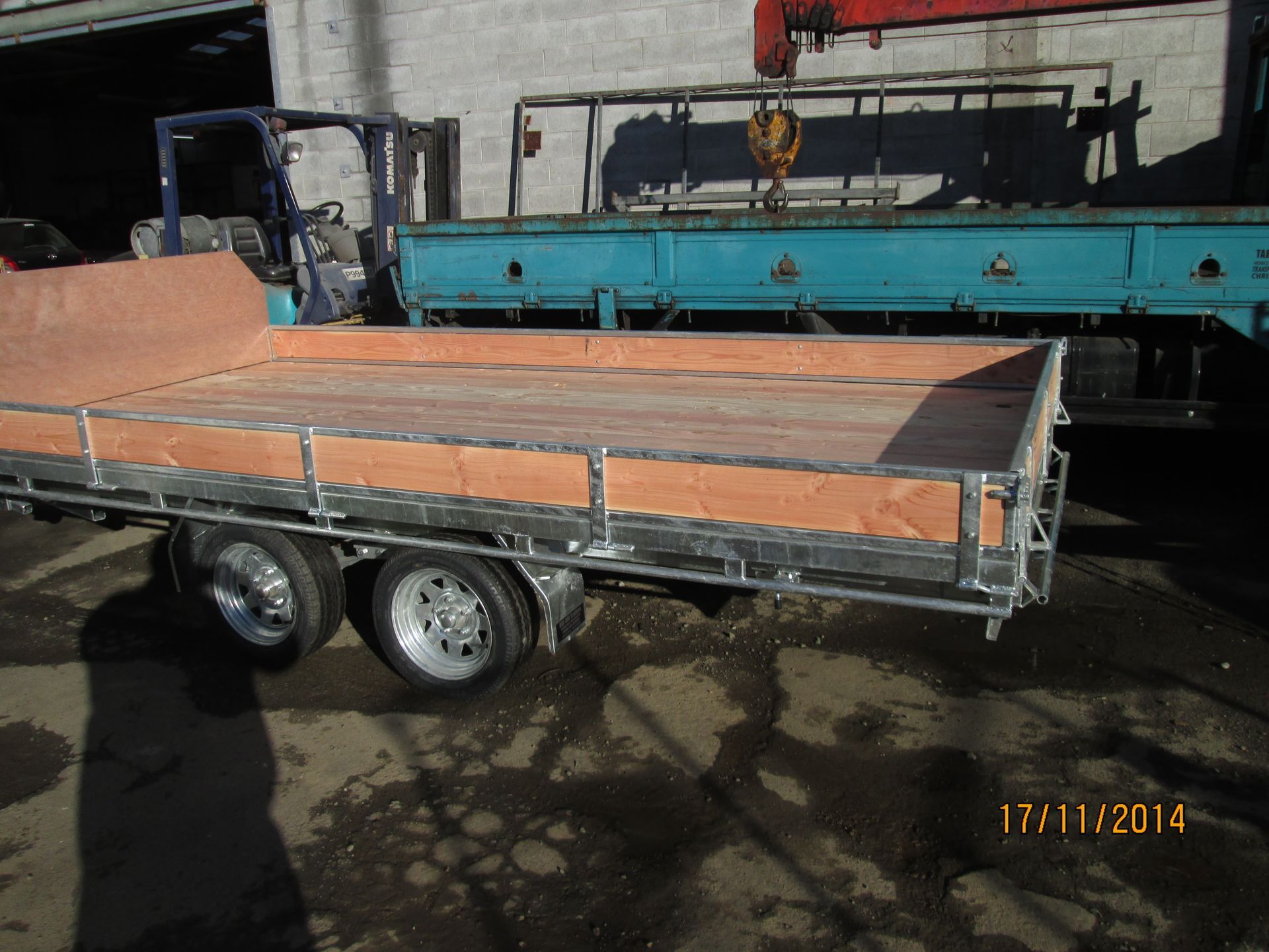 Flatbed trailer with wooden deck, silver frame, parked near blue truck and forklift.