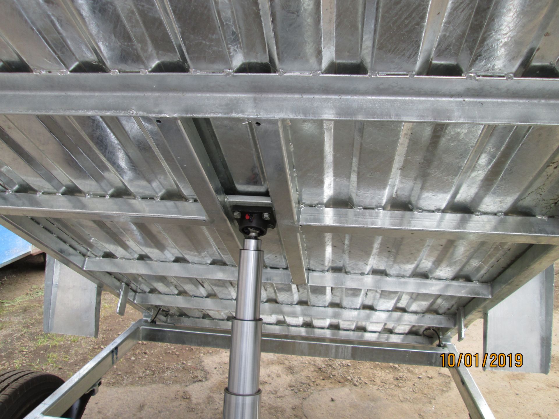 Underside of a metal trailer bed with a hydraulic lift. Silver metal with corrugated design, date stamp on the lower right.