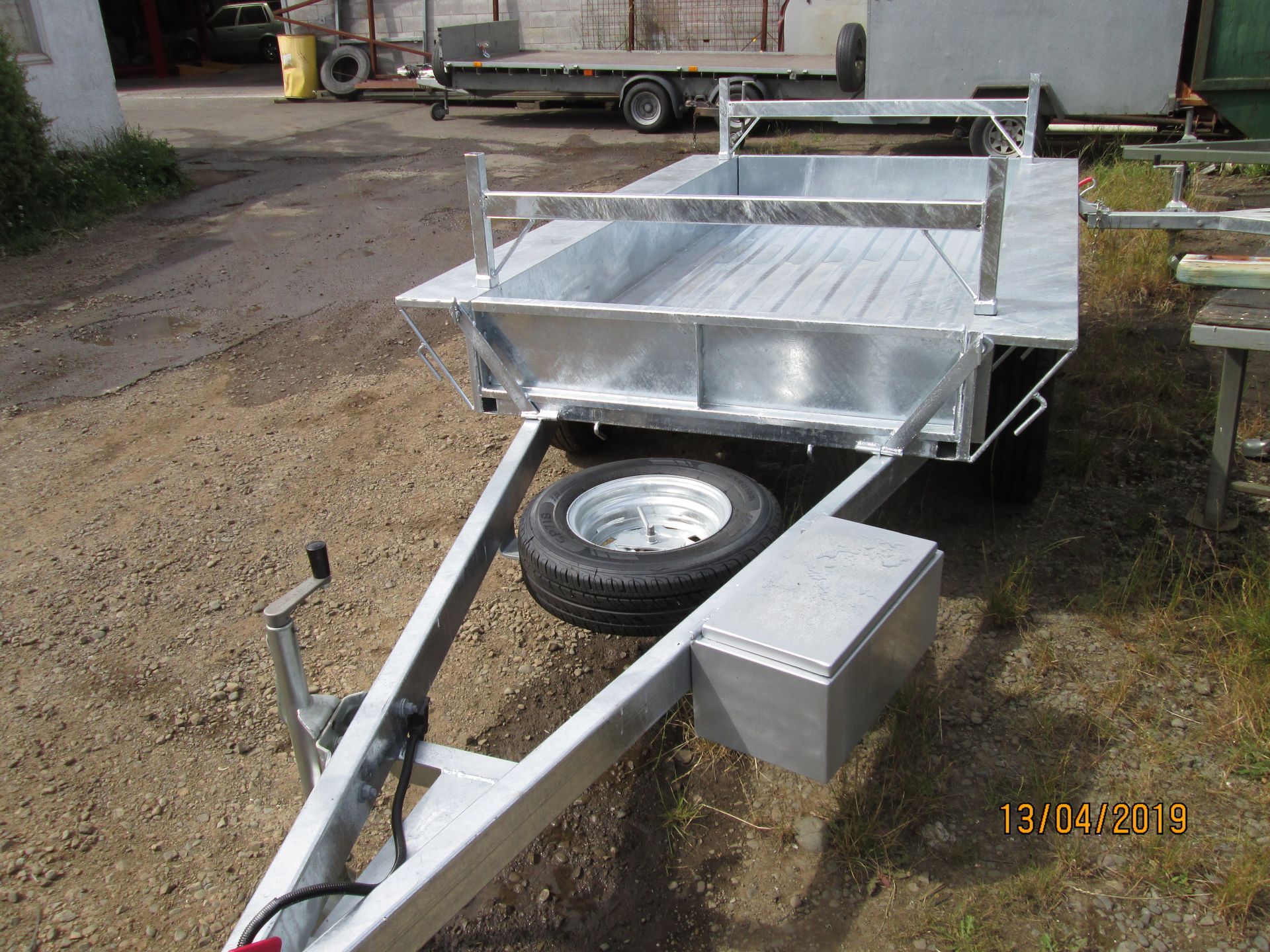 Galvanized metal utility trailer with spare tire and toolbox on gravel.