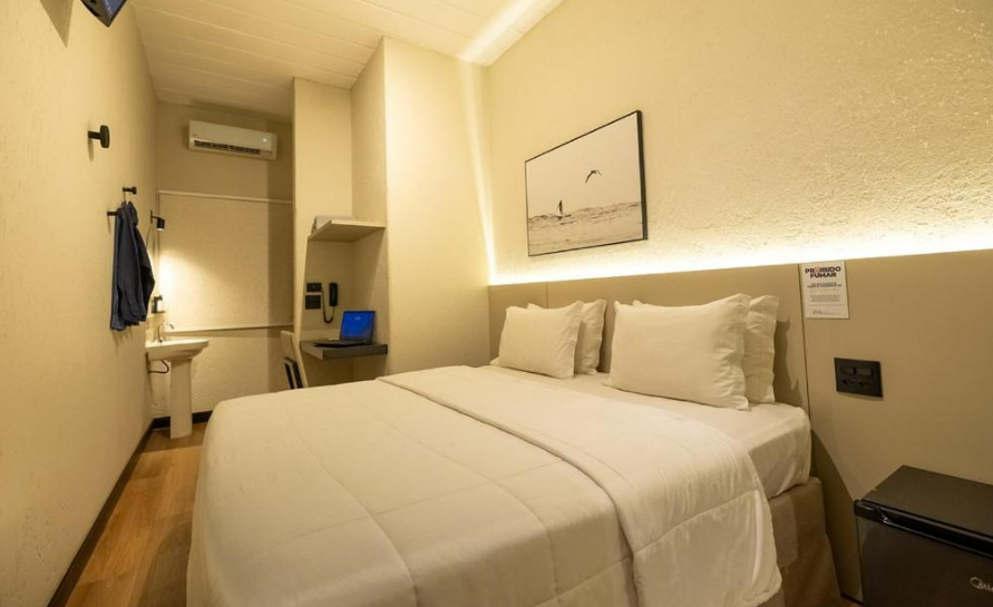 Quarto de hotel com cama, escrivaninha e frigobar. Roupa de cama branca e decoração em tons neutros.