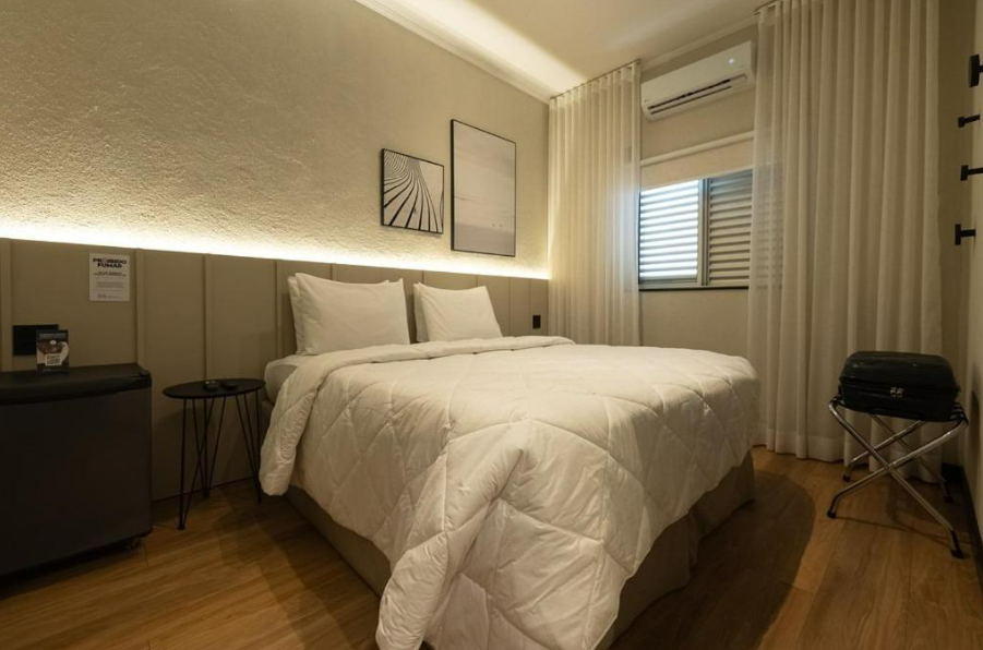 Quarto com cama coberta por um edredom branco. Paredes, obras de arte e cortinas em tons de bege.