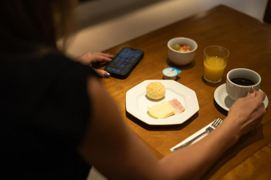 Mulher sentada à mesa com o café da manhã: café, suco de laranja, frutas e um prato com pão, queijo e presunto